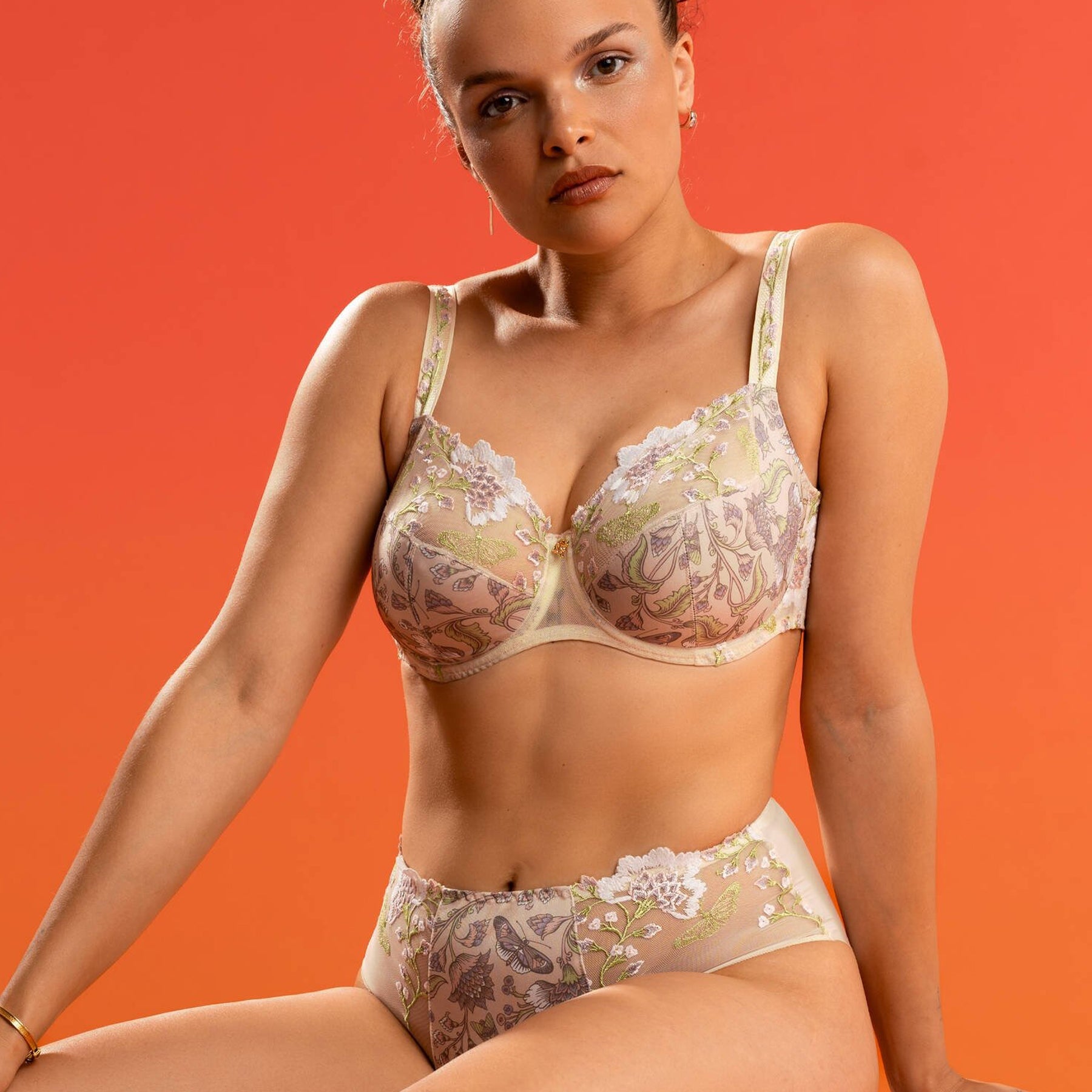 Seated model against a warm orange background wearing a matching embroidered bra and brief set in soft neutral tones with detailed floral design.