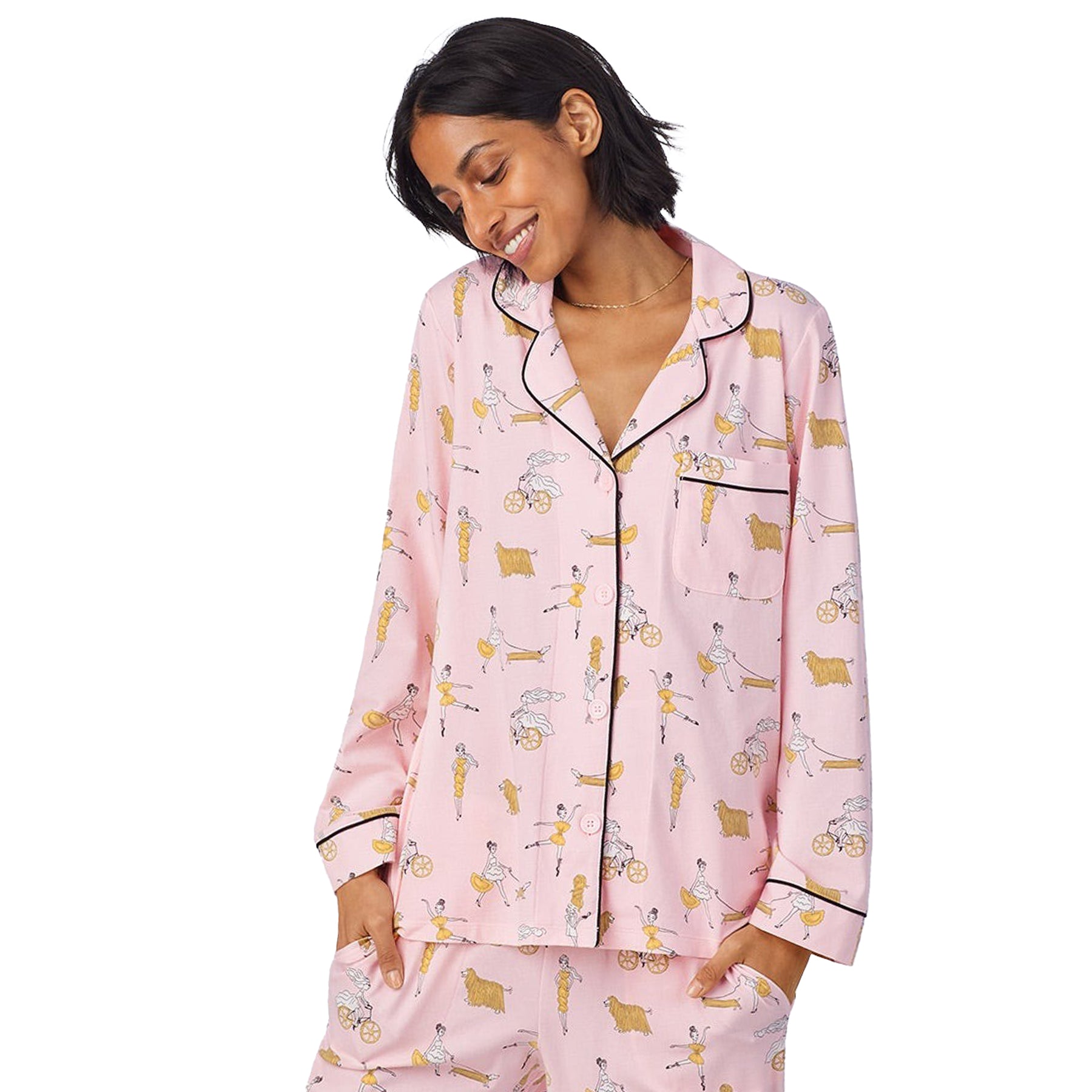 Woman wearing a long-sleeve pink pajama set with black piping and playful illustrated print, smiling with hands in pockets.