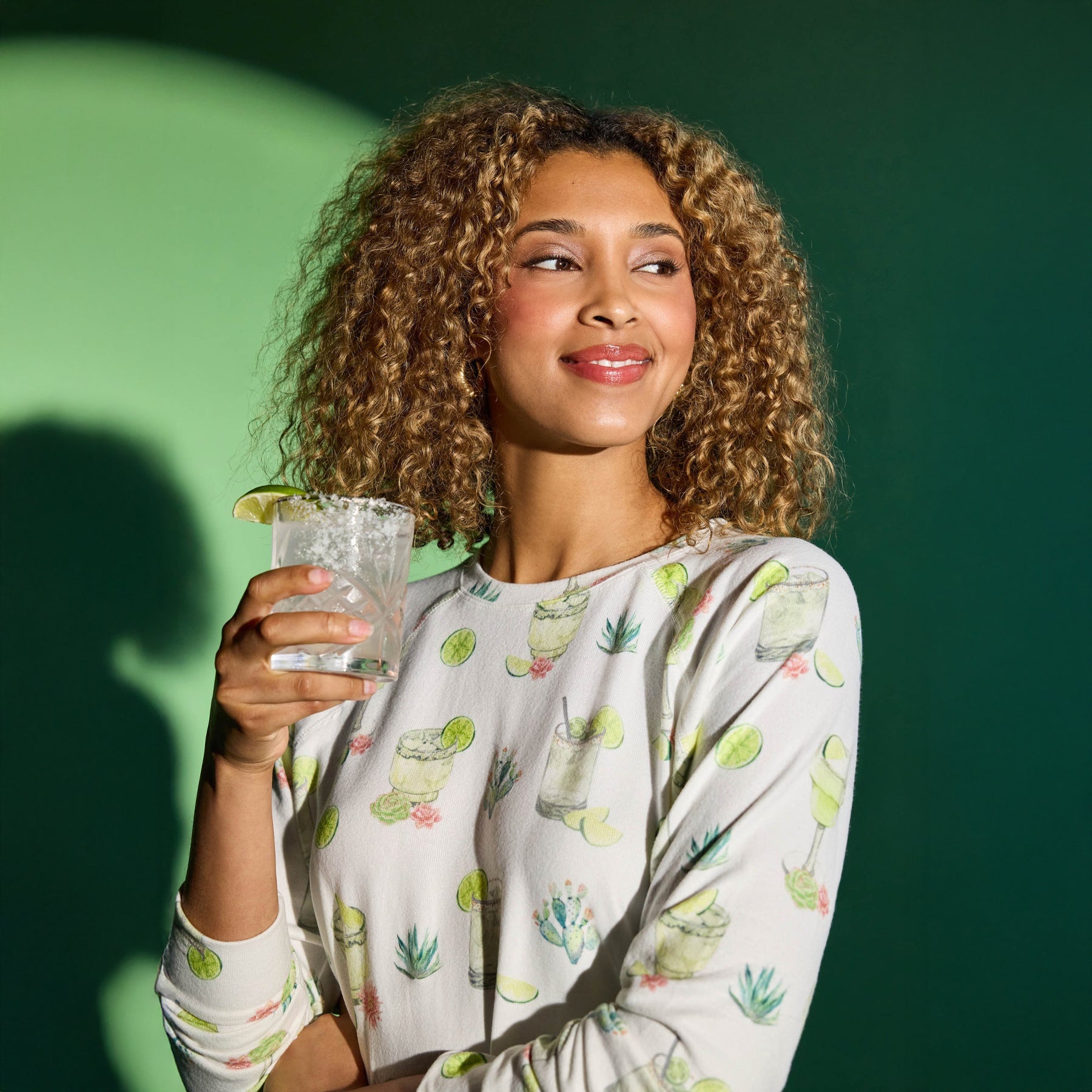 Woman smiling and holding a salt-rimmed margarita glass while wearing a margarita print long-sleeve pajama top.