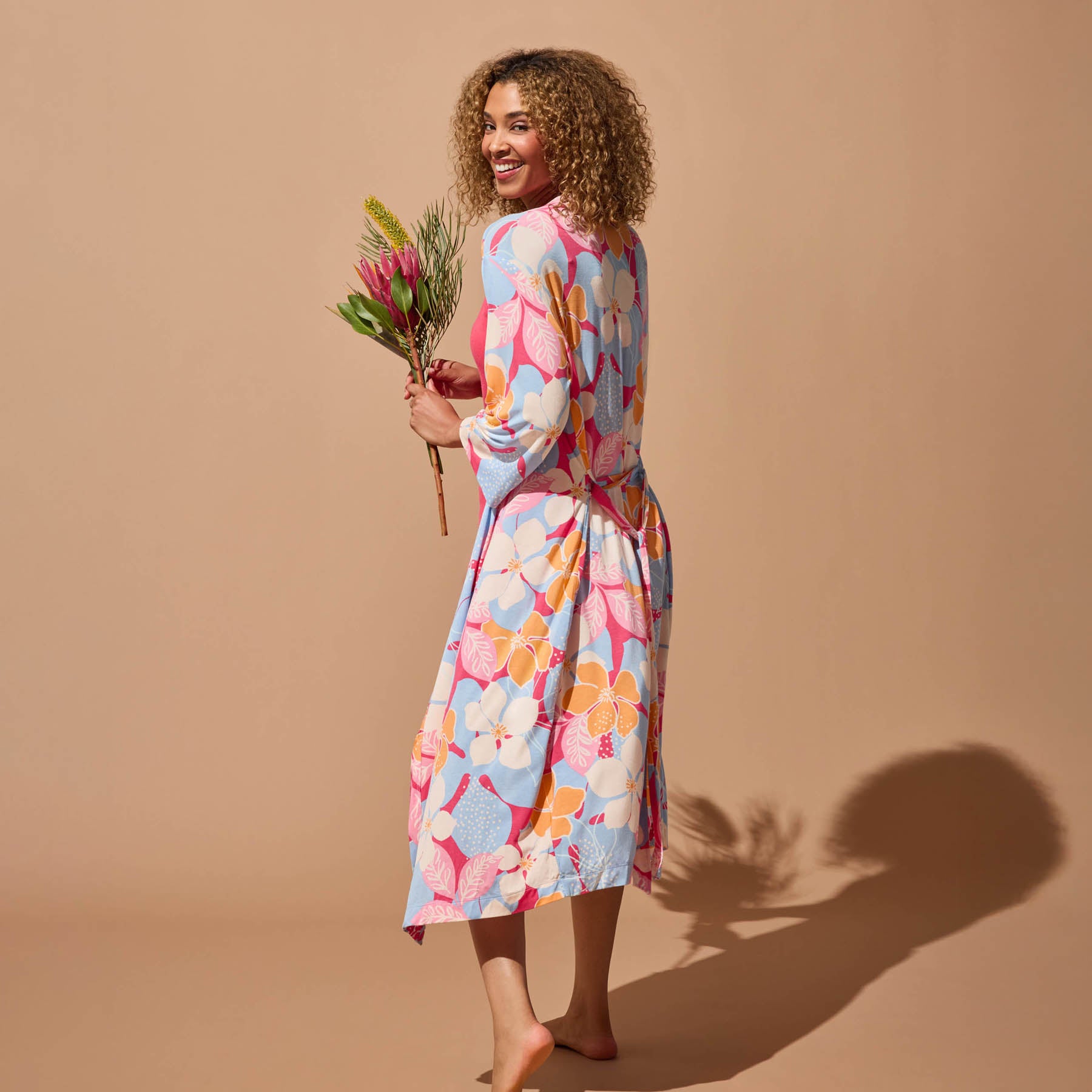 Woman holding flowers, walking barefoot in a colorful floral robe with a tie waist, seen from the back against a tan background.