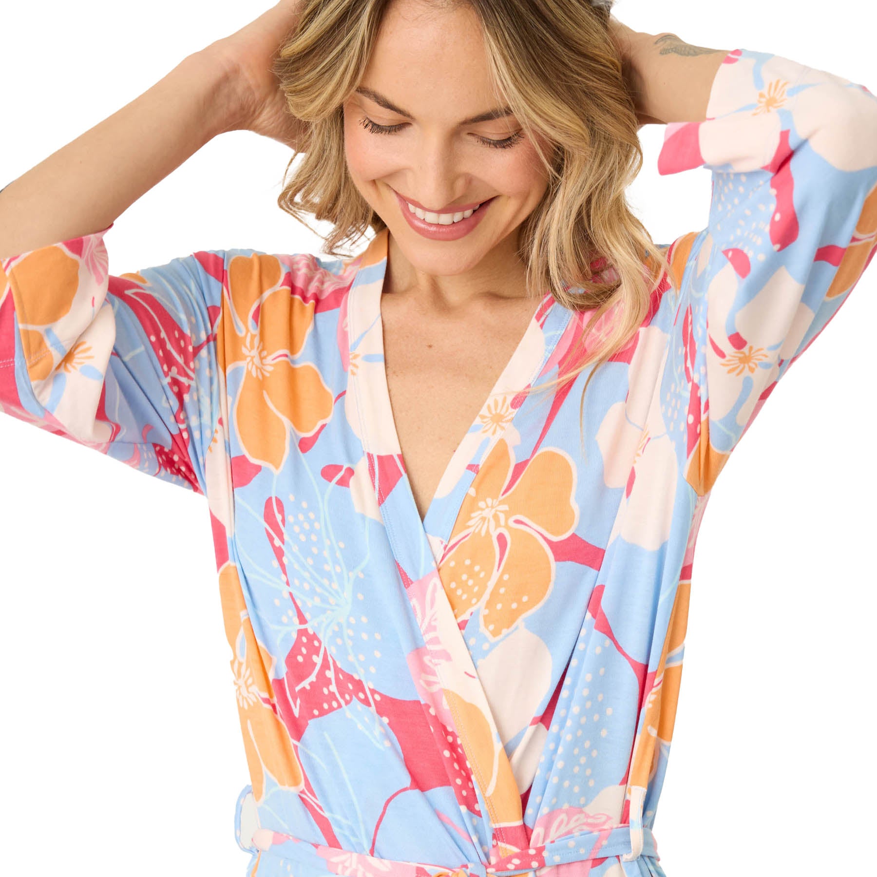 Close-up of a smiling woman adjusting her hair, wearing a wrap robe in a bold tropical floral print with 3/4 sleeves.