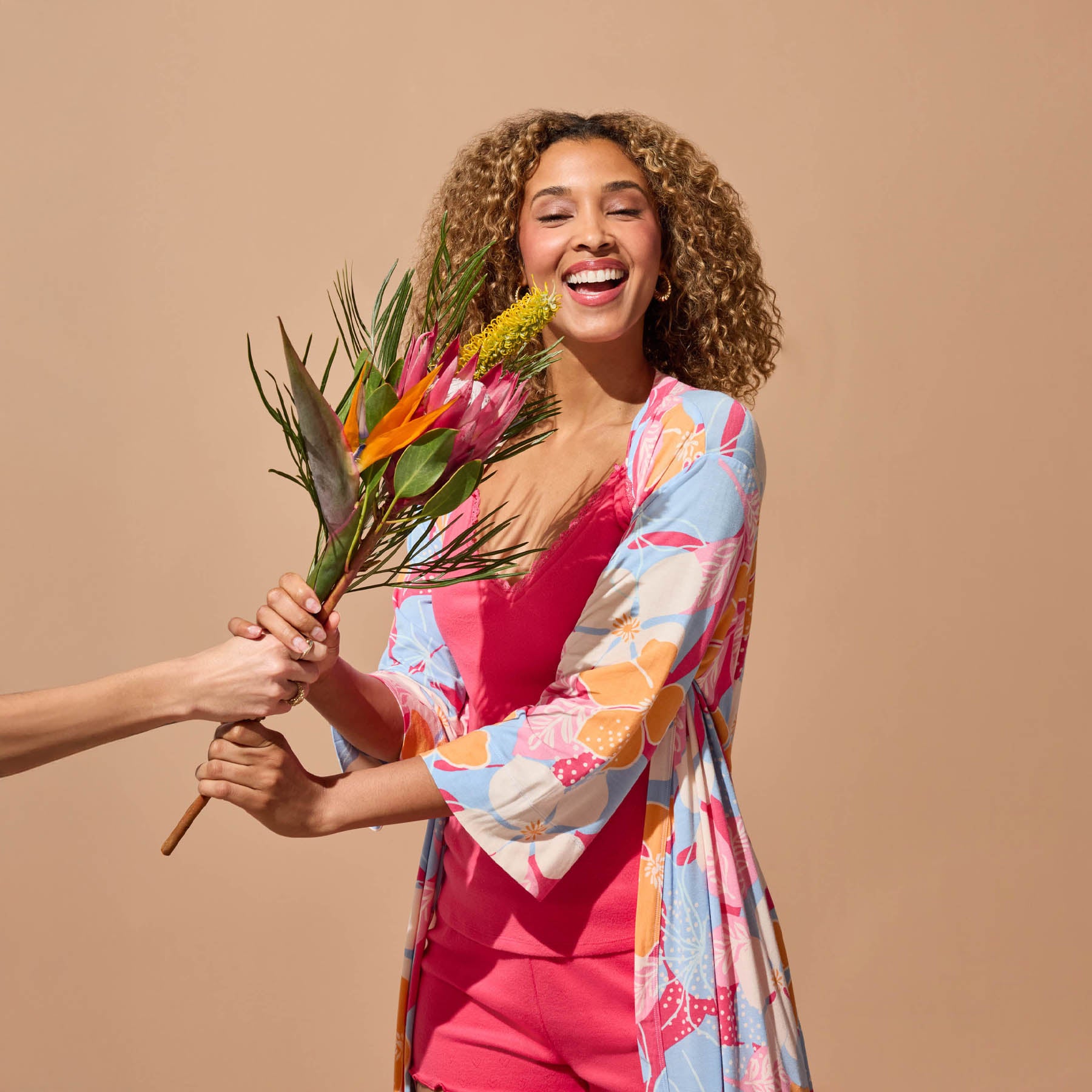 Woman laughing while receiving a bouquet, wearing a floral robe layered over a pink camisole and matching shorts.