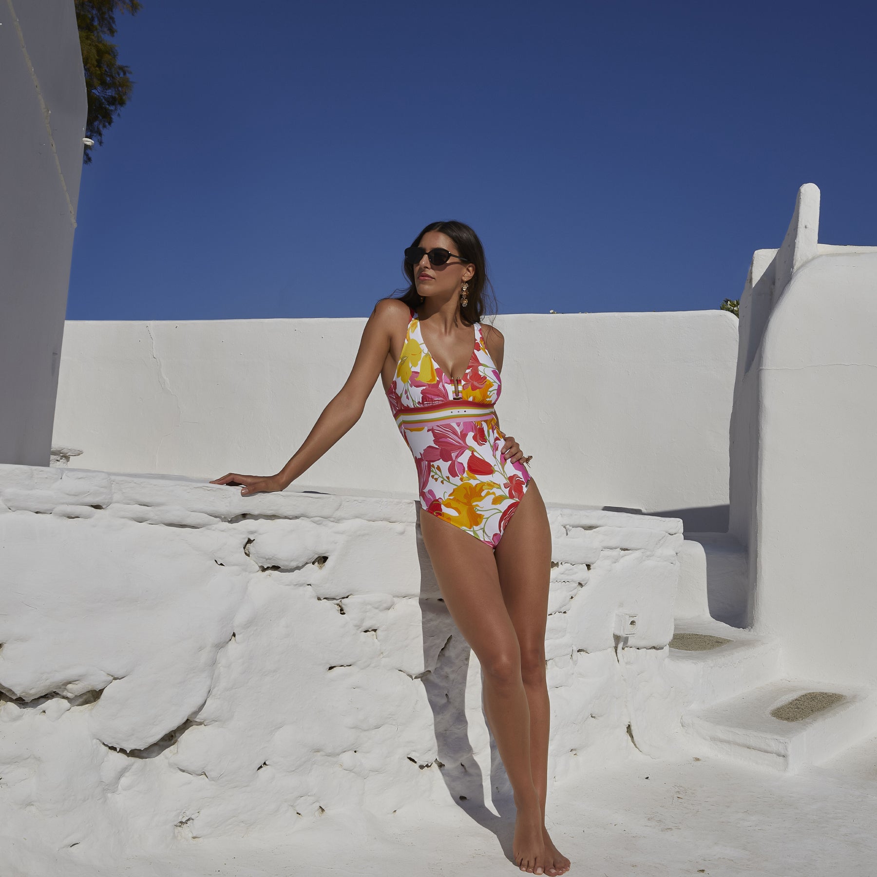 A model wearing a colorful one-piece swimsuit with a bold floral print in pink, red, and yellow hues, featuring a V-neckline and a striped waist detail.