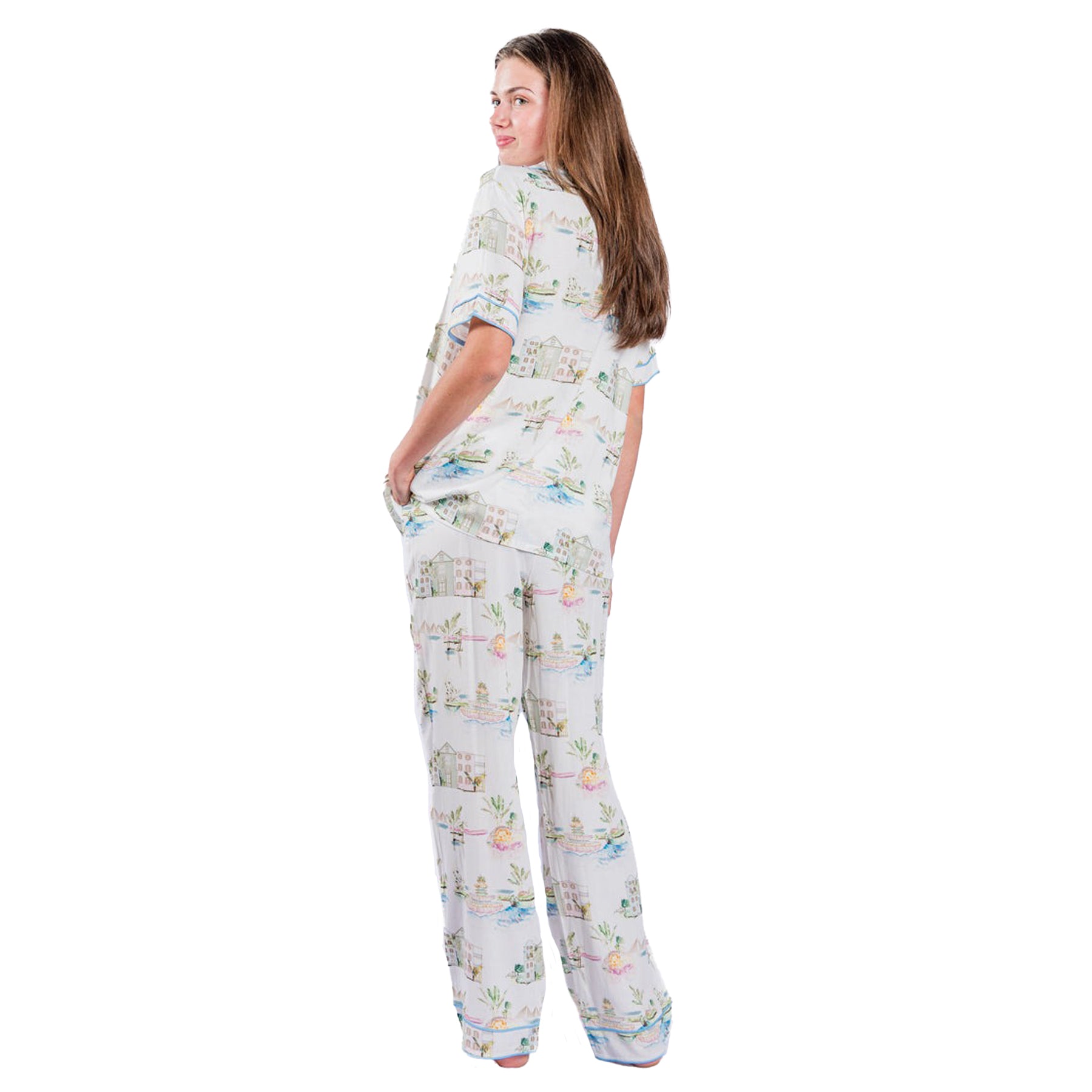 A back-side view of a woman wearing a white PJ SET with a soft, pastel print of cottages, plants, and lakes.