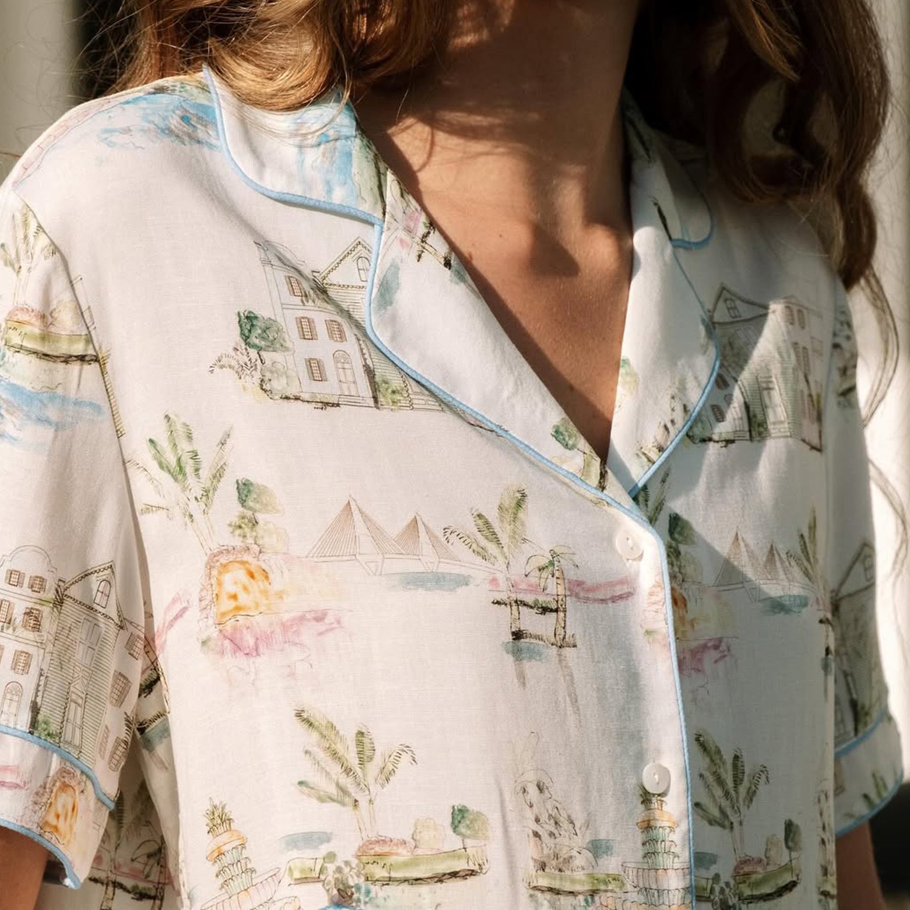 A close-up view of a woman wearing a white PJ SET with a soft, pastel print of cottages, plants, and lakes.