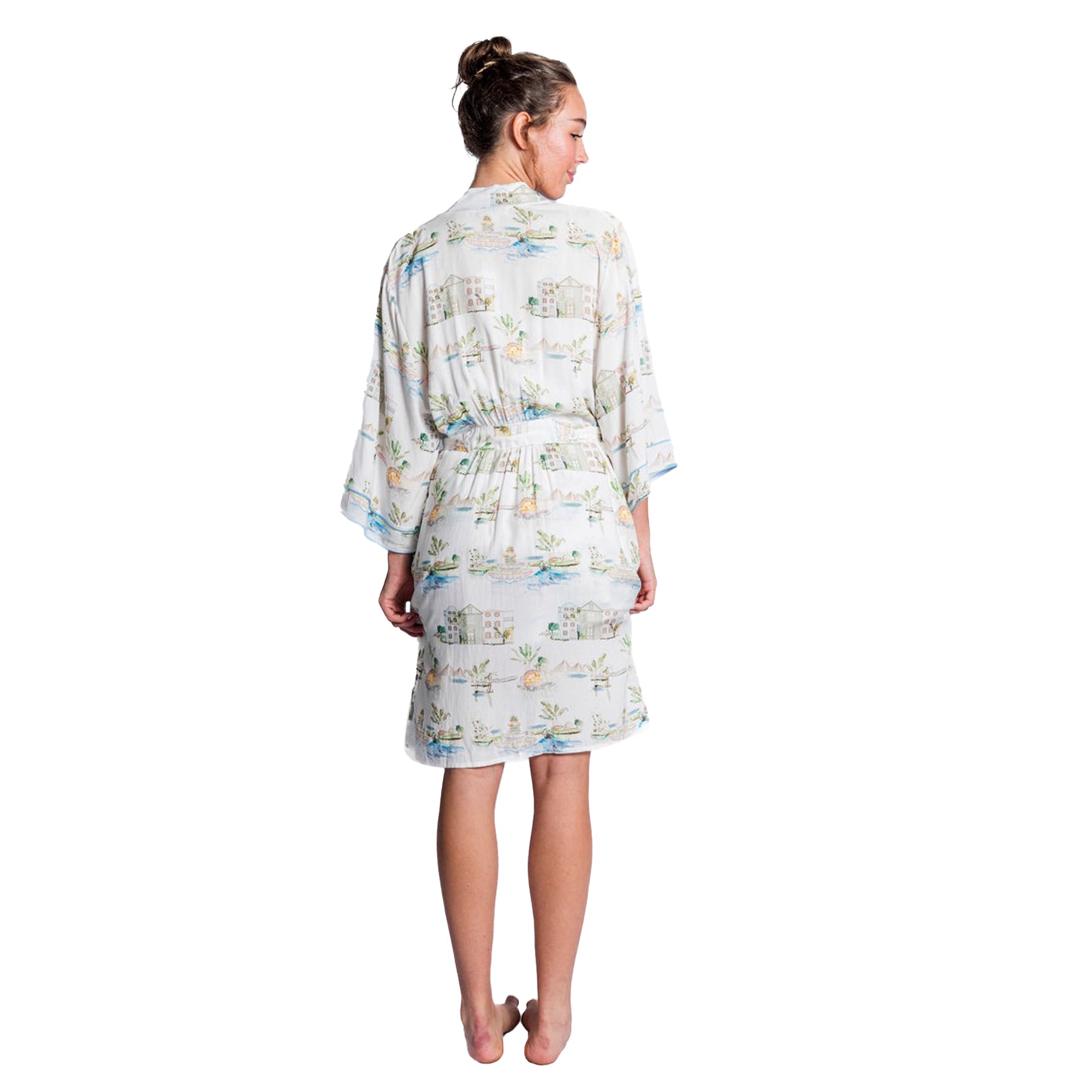 A back view of a woman wearing a white robe with a soft, pastel print of cottages, plants, and lakes. The robe is knee-length with a tie belt at the waist, featuring pockets and a relaxed fit.