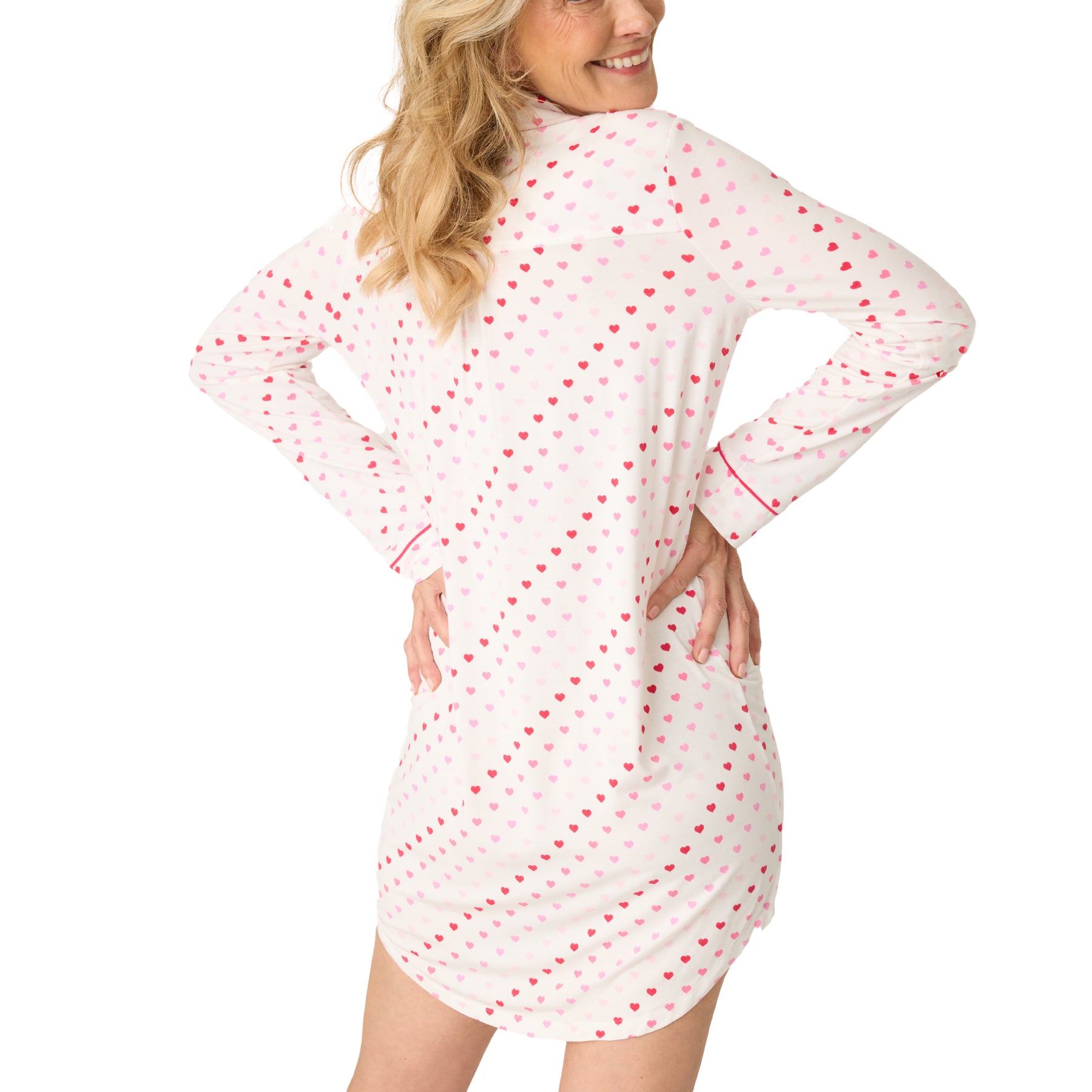 A woman wears a long-sleeve white pajama shirt with a heart print and pink piping, smiling comfortably, view from the back.