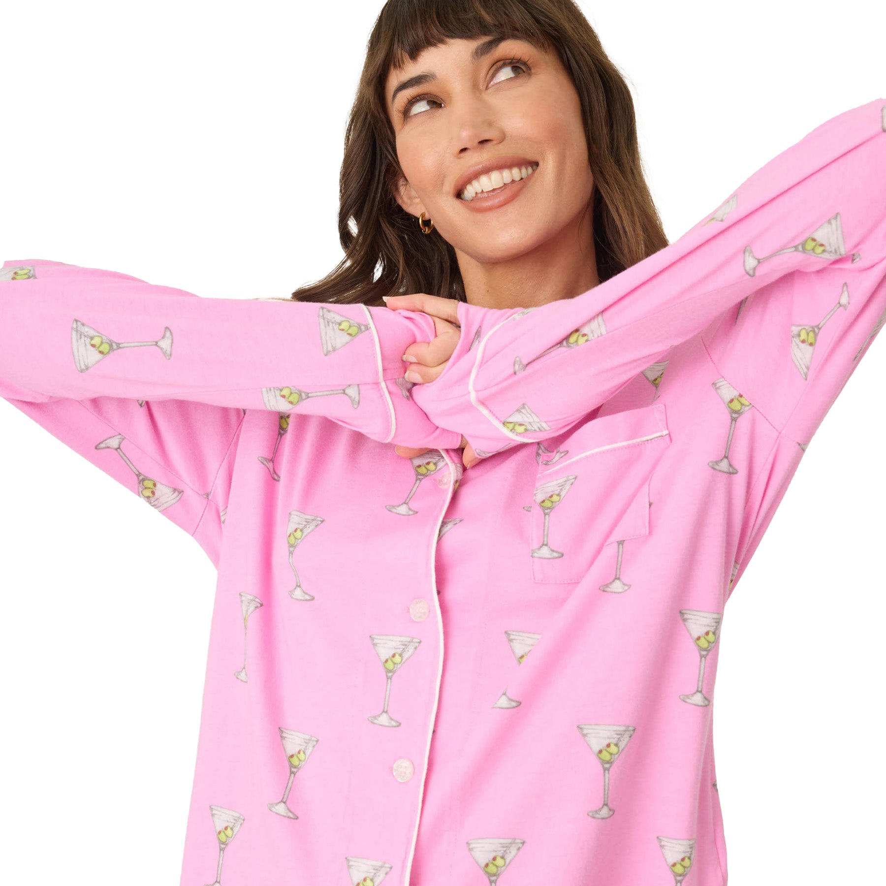 A woman playfully stretching in a pink button-up pajama top with a martini glass print, smiling with her arms raised.