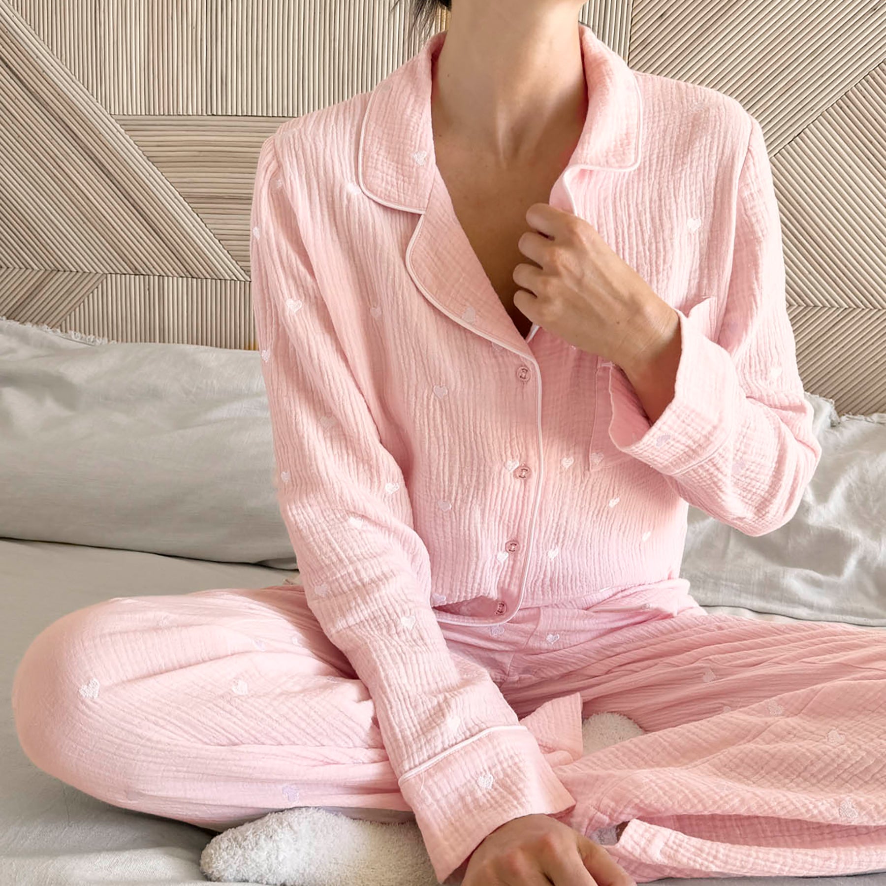 classic button up pyjama set in crinkle cotton in baby pink with mini white embroidered hearts. Matching white  contrast piping on trims. Front view on model sitting.