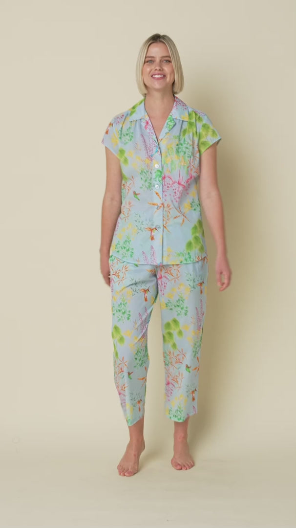 short sleeve button up pajamas with matching capri pants in a multicolour hummingbird print on a blue background. View on model turning to show how it looks.